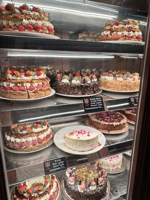 Tủ kính bày các loại bánh kem Anh của AMAM với bảng tên 'British Black Forest Fresh Fruit Cream Bazaar Cake' và 'British Rose Lychee Cream Cake'
