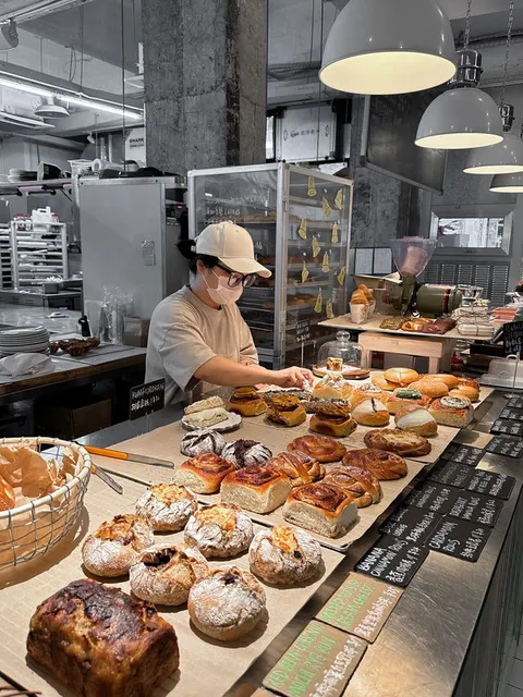 Khay bánh cinnamon roll, cardamom roll và bánh mì tròn xếp đầy quầy, có thẻ tên 'BANANA CINNAMON ROLLS' và 'CARDAMOM ROLLS'