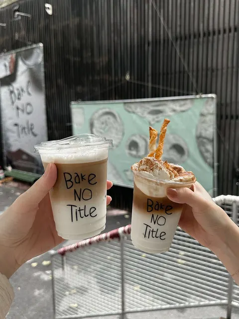 Hai ly cà phê đá in chữ 'Bake NO Title' cầm trên tay, một ly kèm bánh cinnamon roll phủ kem và bánh quế nhỏ