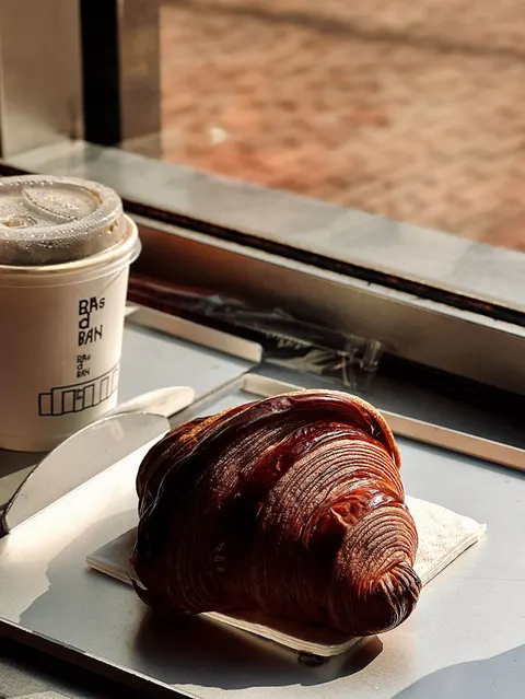 Bánh sừng bò socola bóng vỏ giòn của BAsdBAN đặt cạnh ly cà phê in chữ 'BAs d BAN'