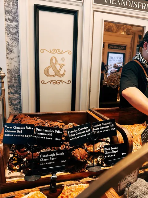 Logo chữ '&' mạ vàng của B&C trên tường, bên dưới là khay bánh cinnamon roll phủ socola với thẻ tên món