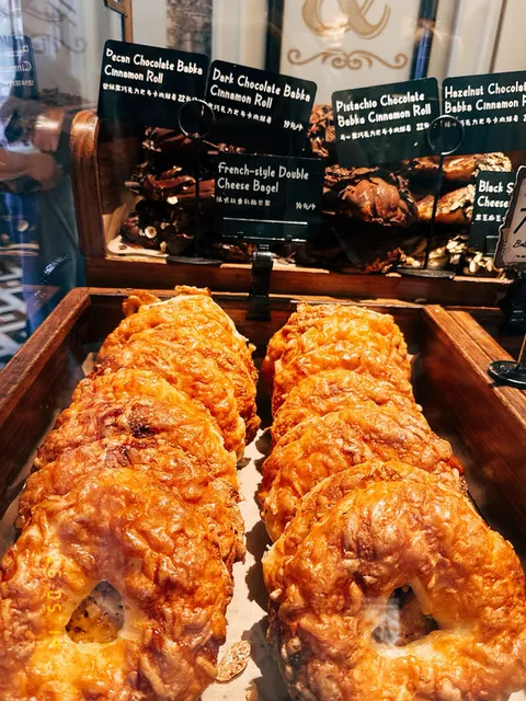 Khay bagel phô mai hai lớp xếp hàng trong tủ kính, bảng tên từng loại bagel/cinnamon roll phía sau