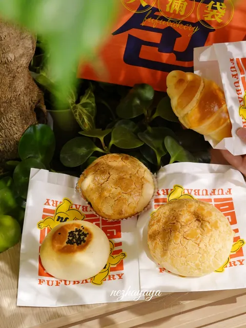 Những chiếc bánh bao mặt nứt giòn đặc trưng (菠萝包) và một chiếc croissant đựng trong túi giấy cam in logo 'FUFUBAKERY'