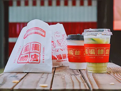Túi bánh giấy trắng in chữ '福福饼店' màu đỏ cùng hai ly trà sữa/cà phê mang thương hiệu 'Fufu Bakery' đặt trên bàn gỗ
