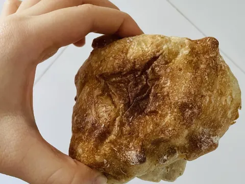 Một tay cầm ổ bánh ciabatta vỏ ngoài giòn rụm, mặt bánh rám nâu đặc trưng của lò nướng thủ công mbd