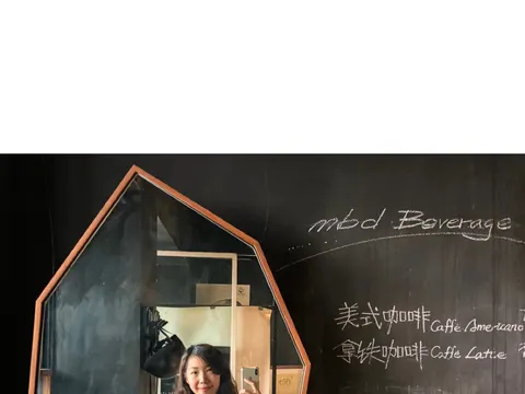 Khách chụp ảnh selfie qua chiếc gương hình tổ ong gắn trên tường đen, bên cạnh là bảng phấn viết tay 'mbd Beverage' với menu cà phê Mỹ và Latte