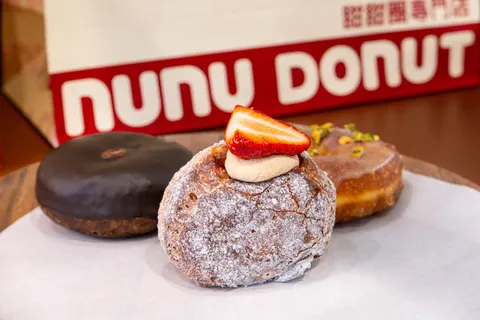 Ba chiếc donut sống đặt trước hộp giấy đỏ-trắng in chữ 'NUNU DONUT', một chiếc phủ đường bột và dâu tươi