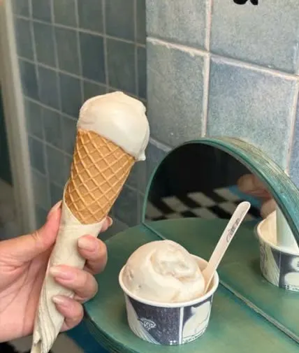 Tay cầm cây kem ốc quế màu trắng ngà cạnh cốc gelato xanh trắng, nền gạch men xanh và gương tròn viền xanh
