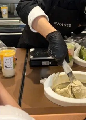 Nhân viên mặc tạp dề CHAGEE, đeo găng đen, múc gelato trà xanh nhạt từ bát đá