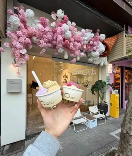 Mặt tiền Osic Gelato màu kem phủ chùm hoa hồng trắng và hồng, tay cầm hai cốc gelato phía trước, ghế xếp ngoài hiên