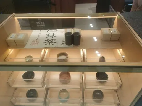 Tủ kính gỗ sáng trưng bày chén trà gốm, hộp trà và tấm giấy chữ 抹茶 trong quán 九十葉 Tân Thiên Địa