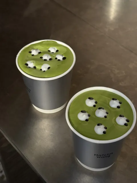 Hai ly Matcha Wang in chữ trên cốc, mặt bọt kem là đàn cừu nhỏ vẽ tay, đặt trên quầy inox