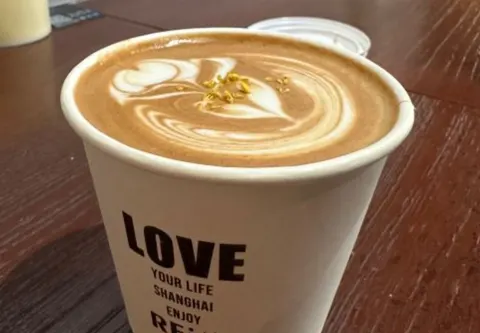 Ly latte hoa quế với cánh hoa quế vàng rắc trên lớp bọt sữa, cốc giấy in chữ REi