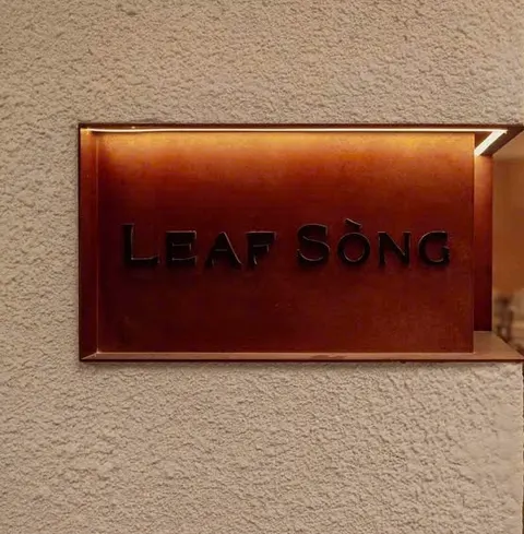 Tấm biển kim loại màu đồng đỏ khắc chữ LEAF SÒNG gắn trên bức tường trát vữa màu kem