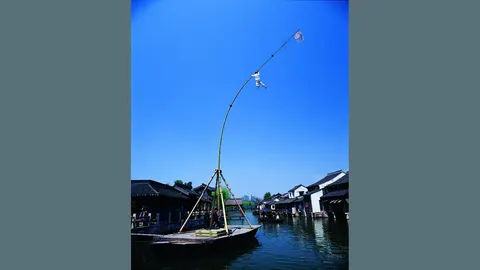 Biểu diễn 'cao can thuyền' ở Đông Sách: nghệ nhân trên cây sào tre cao dựng trên thuyền giữa kênh, hai bên là nhà tường trắng mái ngói đen (ảnh: trang chính thức 乌镇旅游 wuzhen.com.cn)
