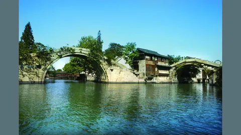 Cảnh Tây Sách (Ô Trấn): hai cây cầu đá vòm bên một thuỷ đình gỗ, mặt nước xanh phản chiếu, trời xanh trong (ảnh: trang chính thức 乌镇旅游 wuzhen.com.cn)