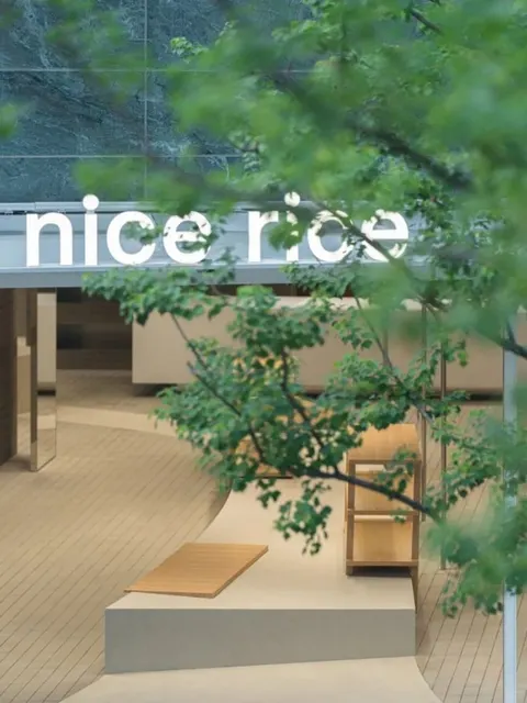 Tiệm nice rice 好饭 ở GATE M 西岸梦中心 nhìn qua tán lá: biển chữ nice rice, bên trong gỗ sáng và đá xám (ảnh: tài khoản tiệm nice rice 西岸梦中心店, qua 空间印象)