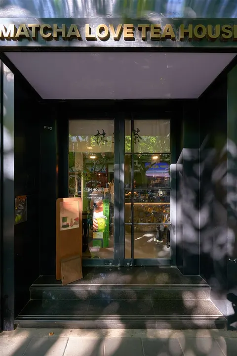 Lối vào Matcha Love Teahouse với biển hiệu đen chữ vàng, cửa kính mở ra khoảng sân nhỏ rợp bóng cây