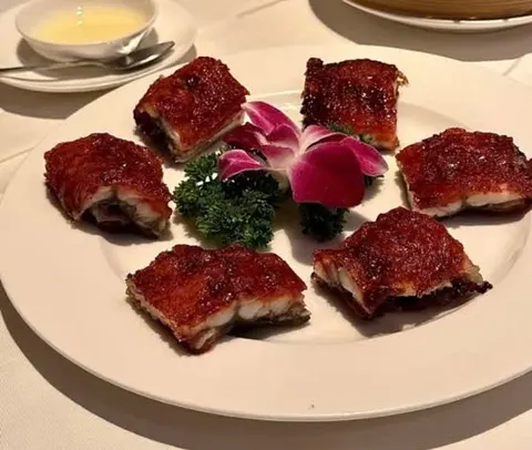 Đĩa heo quay (烧肉) cắt miếng vuông, bì nâu đỏ giòn, giữa đĩa trang trí hoa lan tím và rau xanh (ảnh An gửi)
