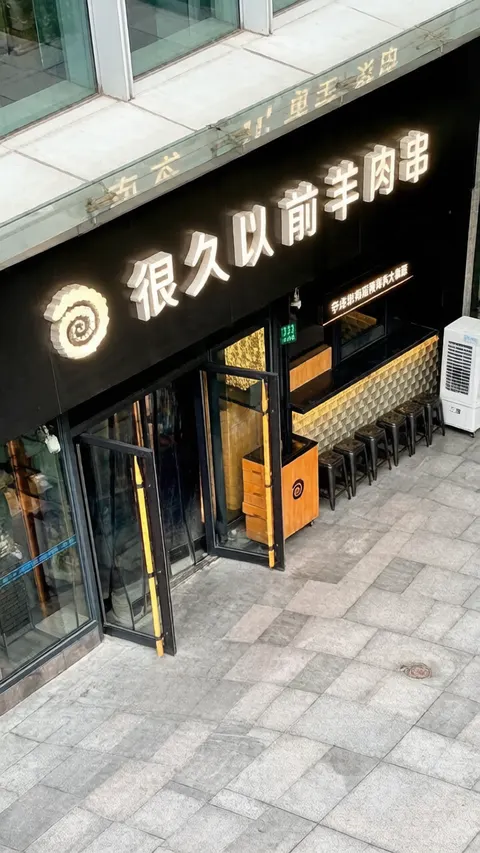 Mặt tiền quán 很久以前羊肉串 chi nhánh Thiên Thược Kiều nhìn từ trên cao: bảng hiệu đen chữ trắng phát sáng, quầy và dãy ghế đẩu