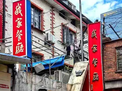 Hai biển dọc màu đỏ chữ vàng 我家餐廳 trên bức tường cũ trong ngõ, cạnh cầu thang sắt và dàn máy lạnh (ảnh trang Trip.com của 我家餐厅 华山路店)