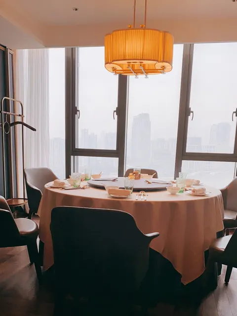Bàn tròn trải khăn màu đào với mâm xoay, đèn thả trụ màu cam, cửa kính lớn nhìn ra đường chân trời thành phố