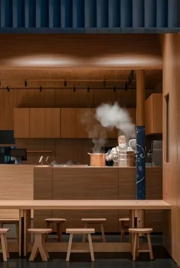 Quầy pha chế mở ốp gỗ sáng màu, dải lam xanh đậm phía trên, nhân viên đứng nấu bên nồi đồng bốc hơi