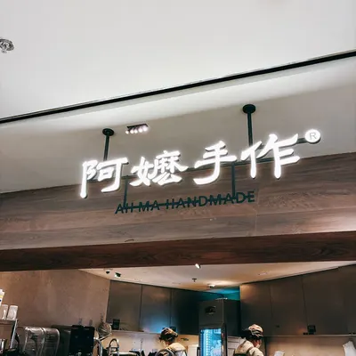 Biển hiệu chữ nổi sáng '阿嬷手作 AH MA HANDMADE' phía trên quầy pha chế gỗ tối màu, hai nhân viên đang làm việc bên dưới