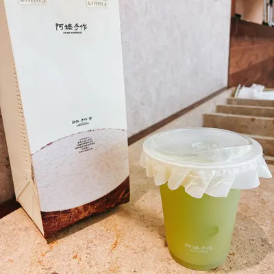 Cốc trà màu vàng xanh in logo 阿嬷手作 đặt cạnh túi giấy thương hiệu, phía sau là quầy pha chế