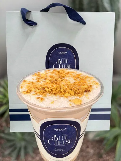 Tay cầm cốc trà phủ kem phô mai của 椿田熟成茶行 (CHUNTIAN), cạnh túi giấy xanh nhạt in logo quán, ánh sáng ấm phía sau