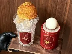 Ly trà đá phủ kem phô mai đặt cạnh lon trà thiếc đỏ trên quầy gỗ tối màu, decor phục cổ kiểu Anh của quán