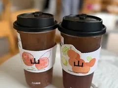Hai cốc trà màu nâu cam có hình quả hồng và chữ 'Shan' trên nhãn, đặt cạnh nhau trên bàn trắng