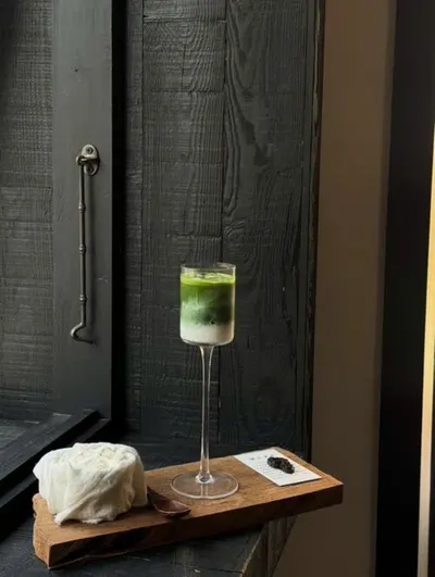 Ly trà matcha chân cao và miếng phô mai trên khay gỗ cạnh cửa sổ (ảnh An gửi, 08/10)